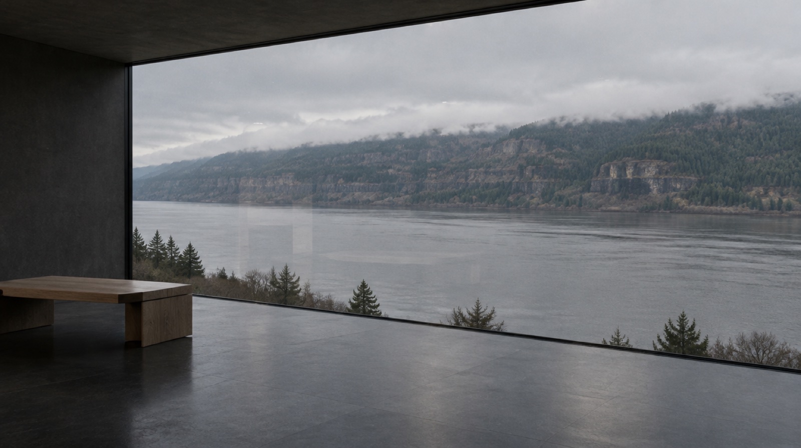 A dark room with a basalt floor and oak bench, looking through a floor-to-ceiling frameless glass wall at a wide grey river and fir-covered cliffs.